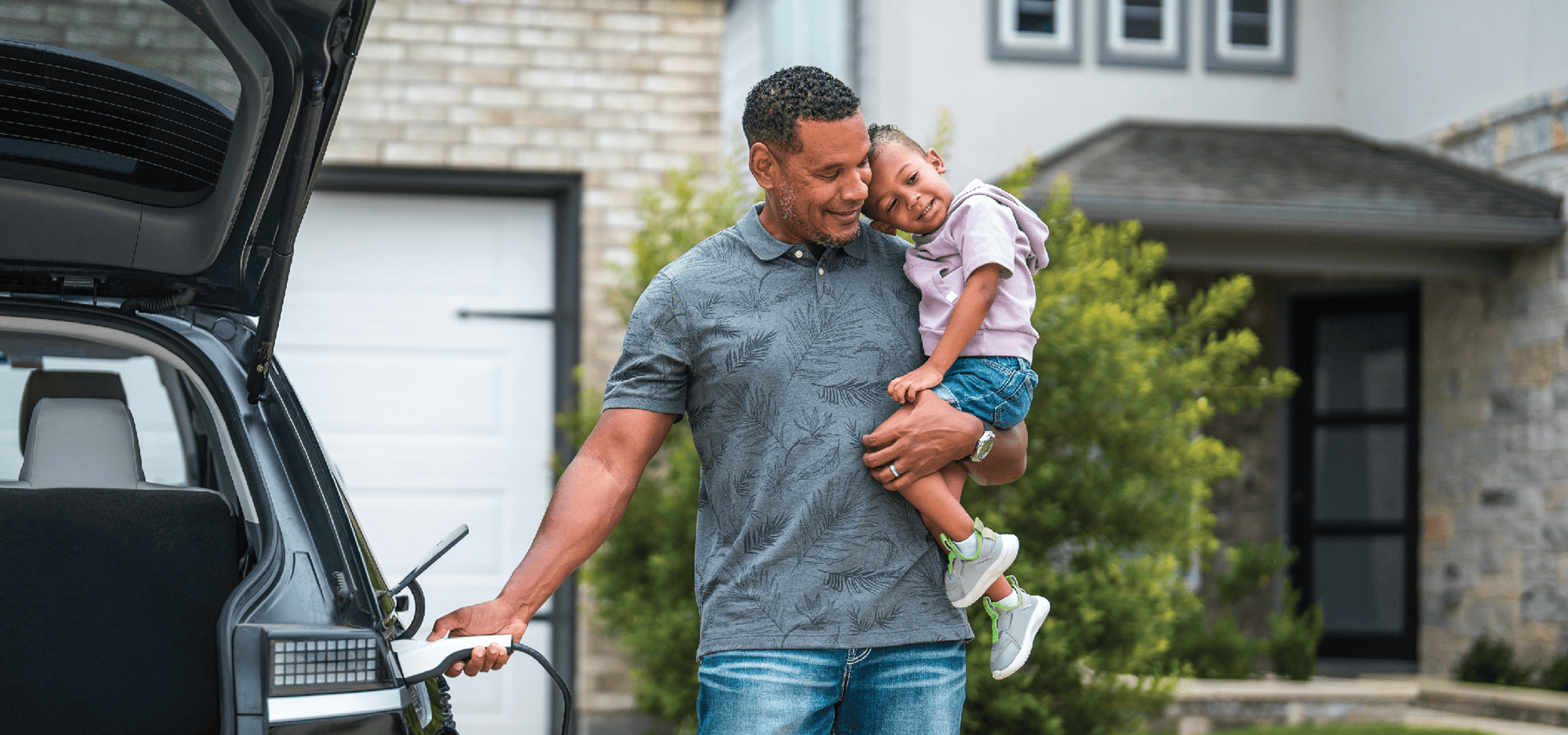 Father holding daughter while plugging charger into electric car in front of their house.